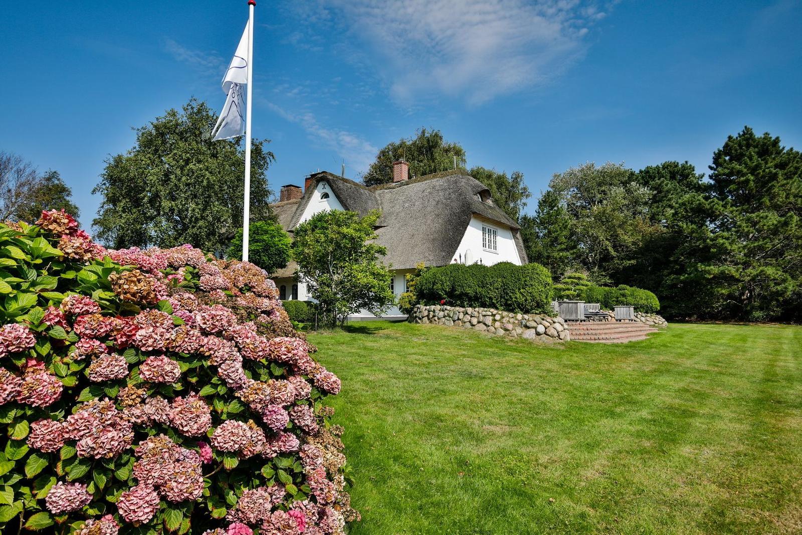 House with thatched roof, green lawn, and flower hedge under blue sky.