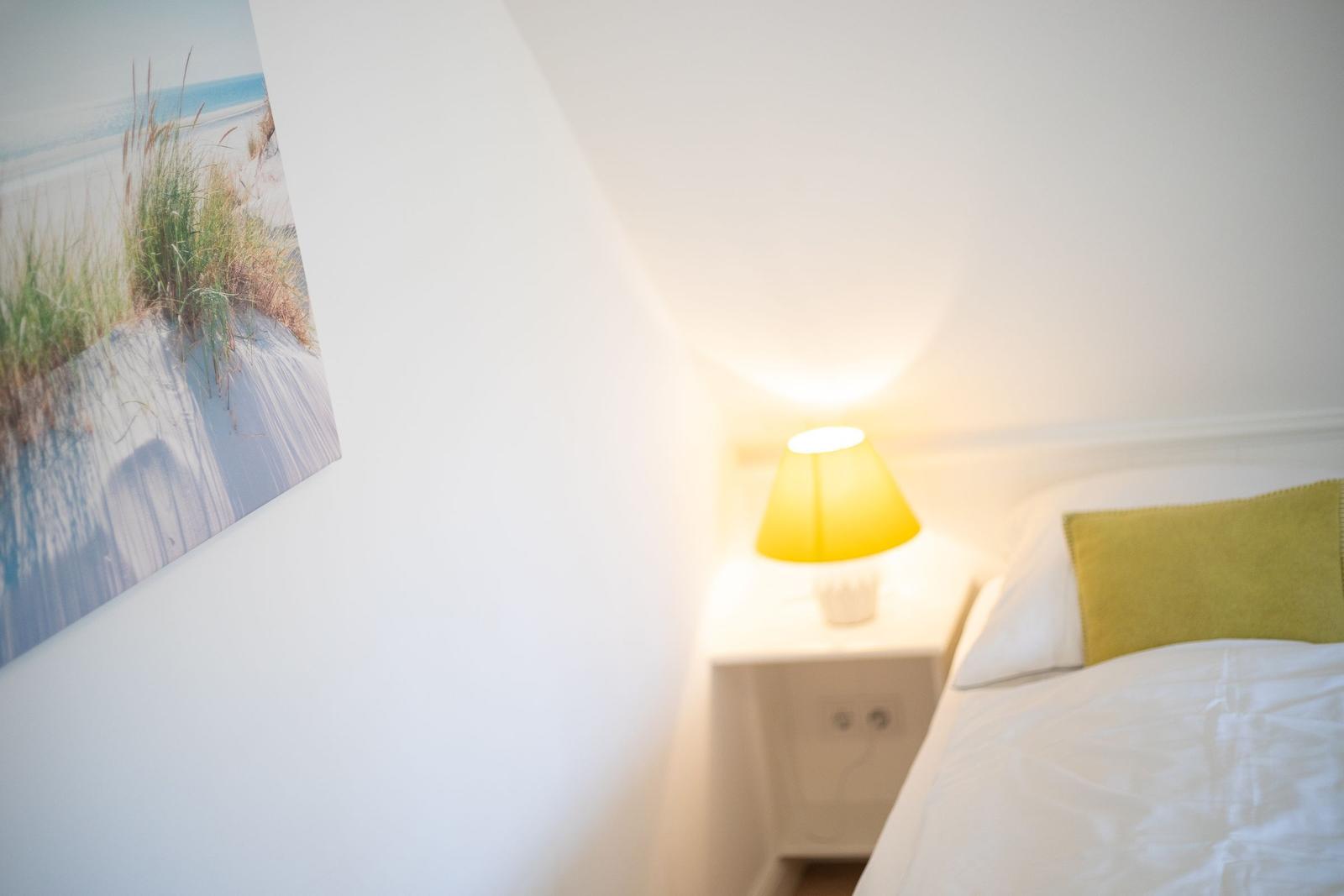 Schlafzimmer mit Bett, Nachttisch und Lampe. Wandbild zeigt Strand.