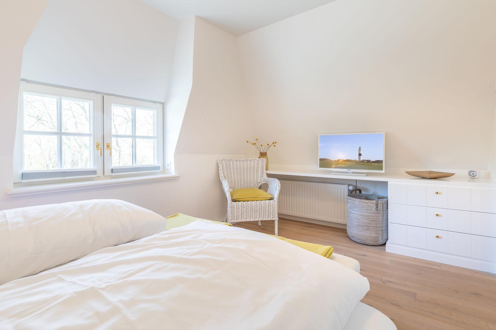 Schlafzimmer mit Bett, Fenster, Stuhl und Fernseher an der Wand.