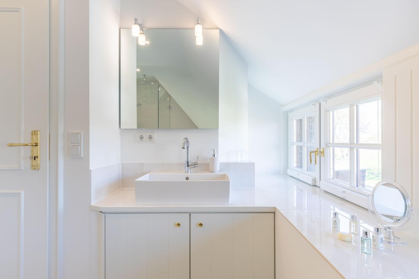 Badezimmer mit Waschbecken, Spiegel und Fenster. Weiße Fliesen und goldene Griffel.