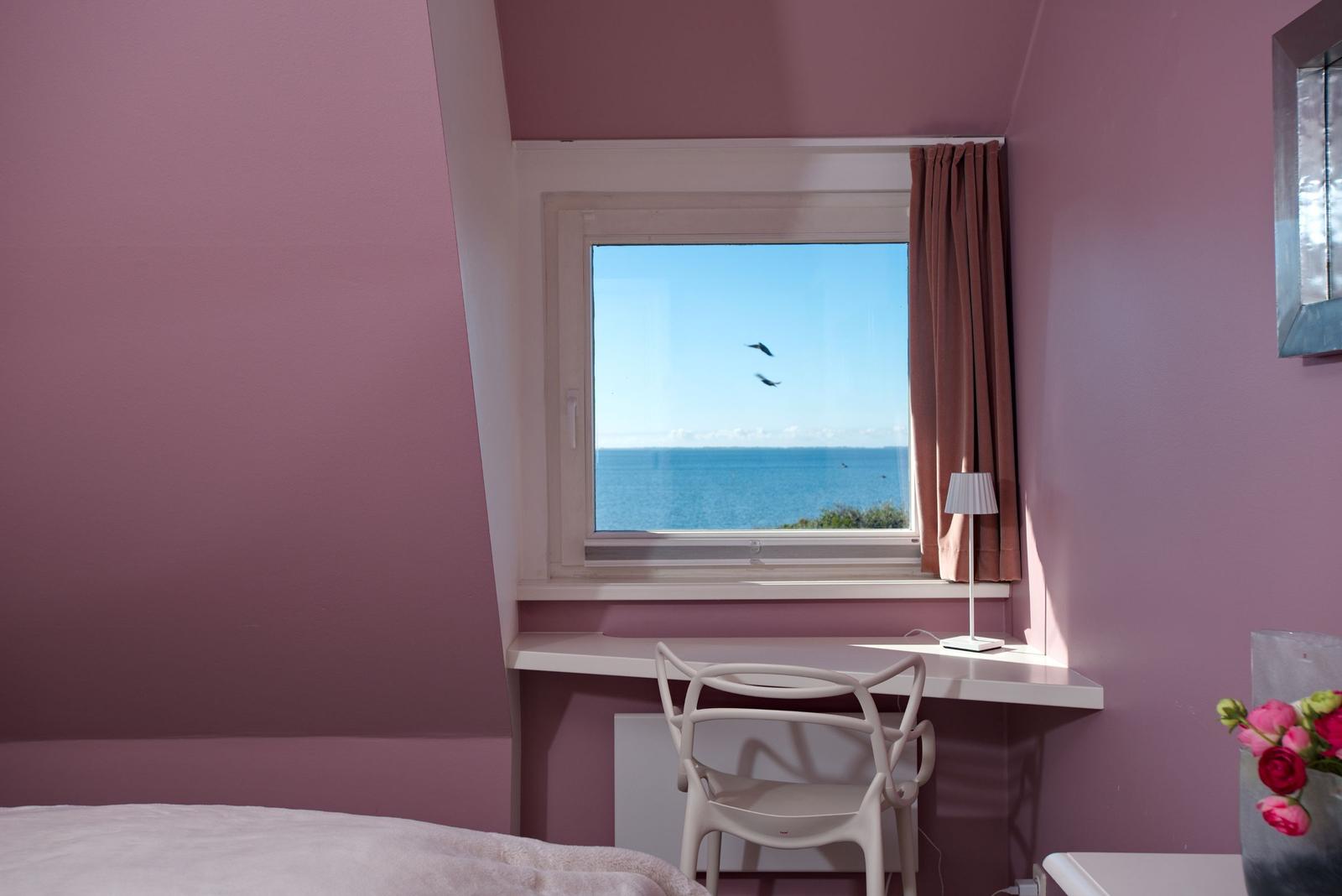 Ein Fenster mit Meerblick, Schreibtisch und Stuhl in einem Raum mit rosa Wänden.