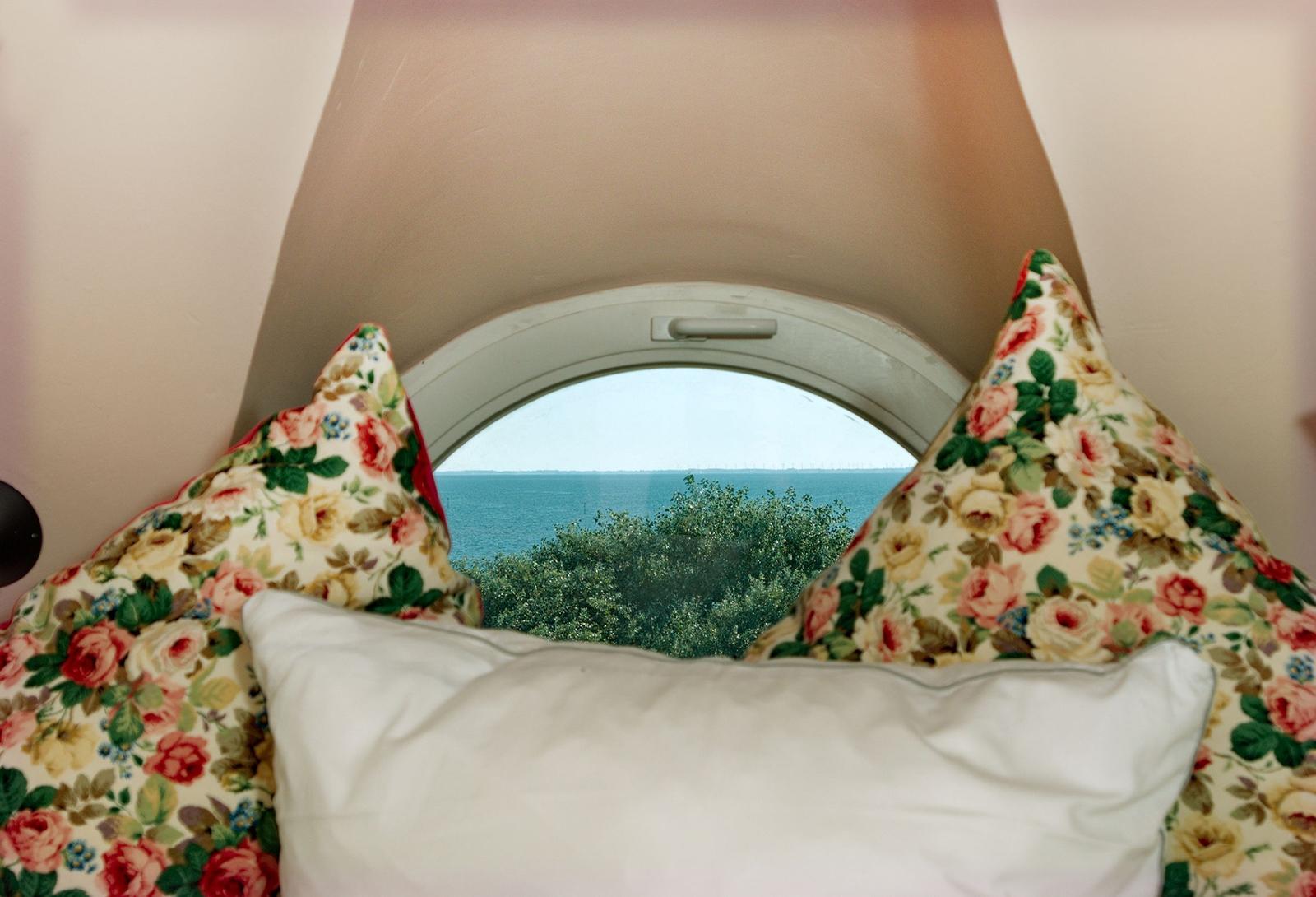 Rundes Fenster mit Blick auf Meer und Bäume, zwei Kissen mit Blumenmuster vorne