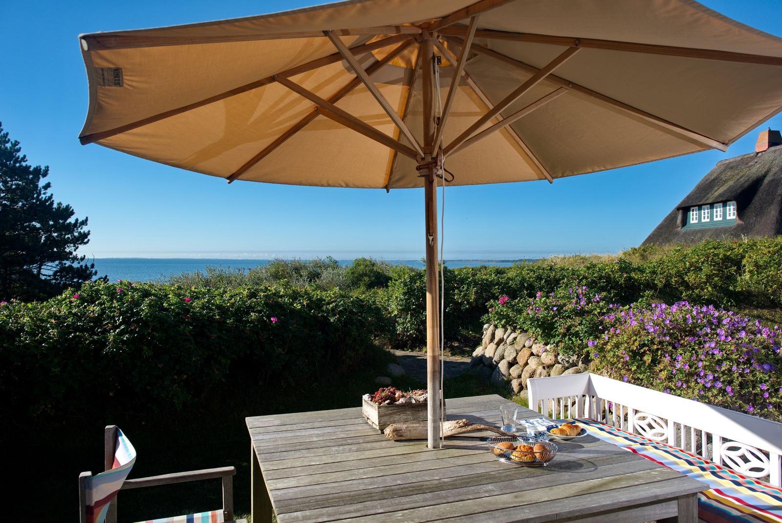 Terrasse mit Tisch, Sonnenschirm und Meerblick. Blumenbeete und ein traditionelles Haus sind sichtbar.