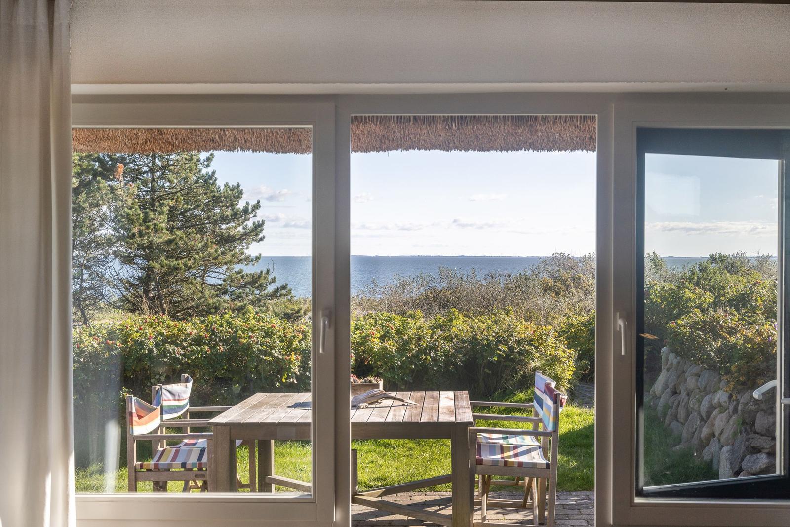Durch die Glastüren blickt man auf einen Garten mit Tisch und Stühlen. Im Hintergrund ist das Meer zu sehen.