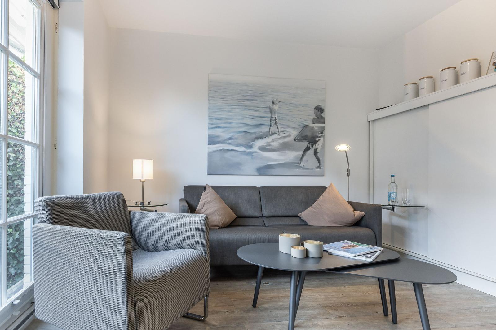 Wohnzimmer mit grauem Sofa, Sessel und runden Tisch. Wandbild und Fenster sichtbar.