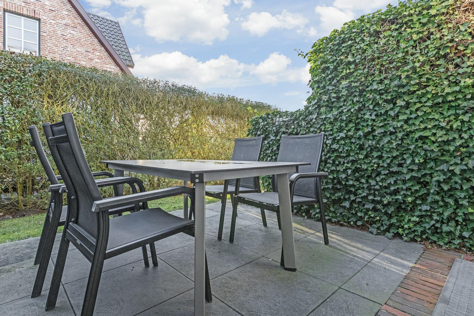 Terrasse mit Tisch und vier Stühlen, umgeben von grünen Hecken und einem Haus.