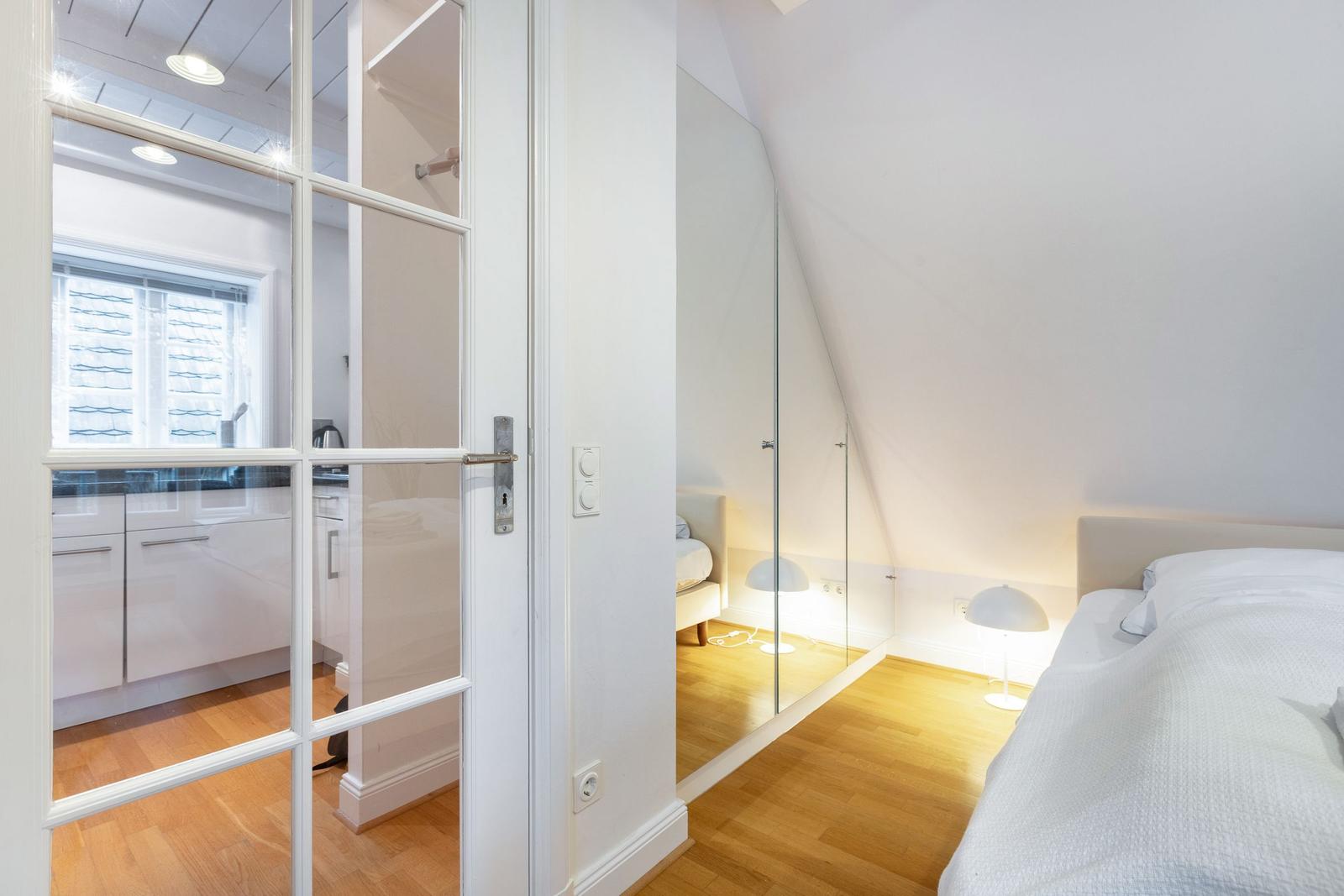 Schlafzimmer mit Dachschrägen, Bett, Lampe und Glaswand. Tür führt in Küche mit Fenster.