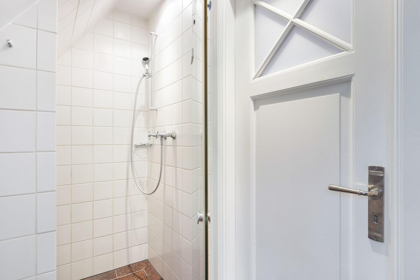 Dusche mit Duschbrause und weißen Fliesen. Tür mit Metallgriff.