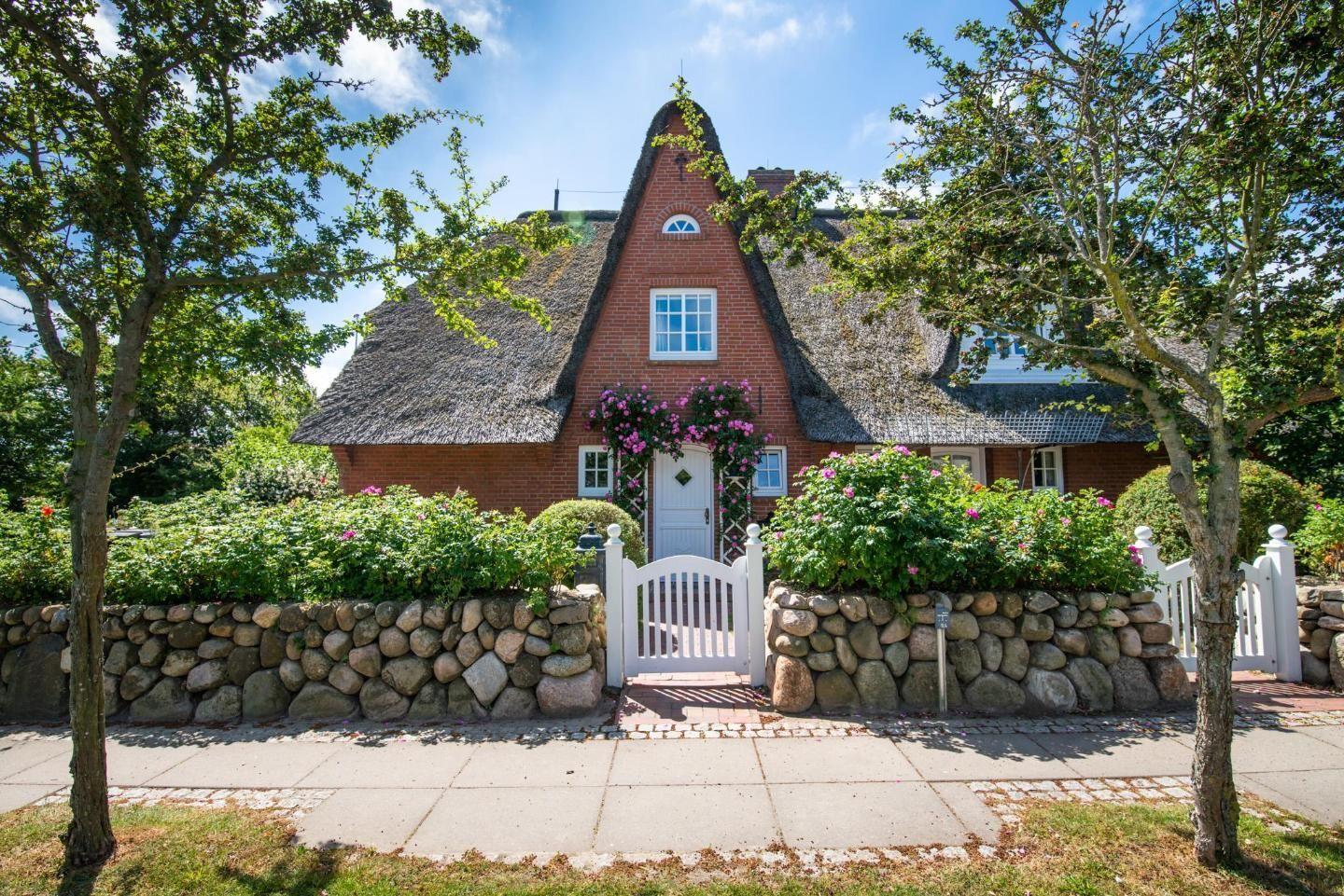 Red brick house with thatched roof and white door. Garden with stone wall and flowers.