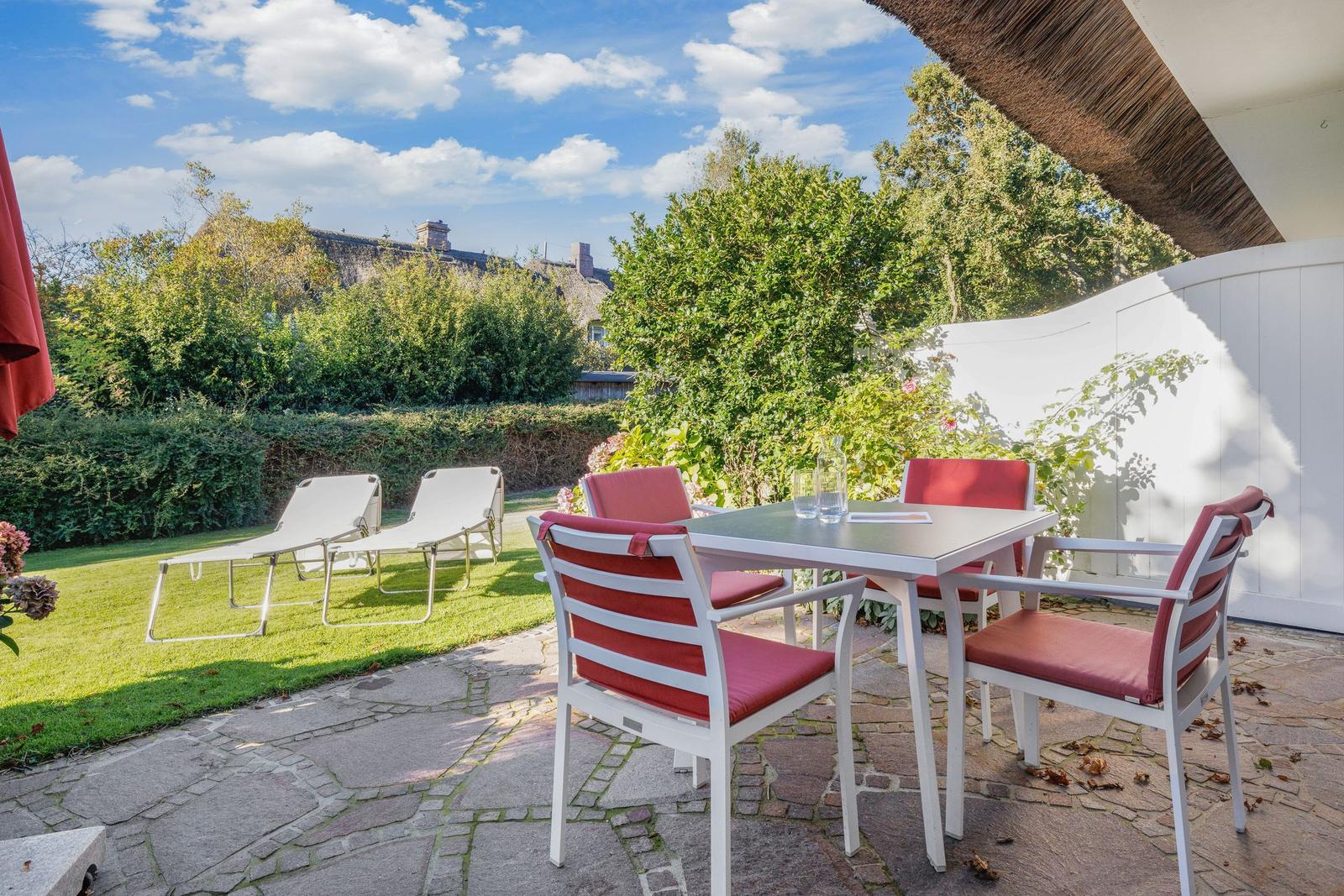 Terrasse mit Tisch, Stühlen und Liegen im Garten. Hintergrund: grüne Hecke und Himmel.