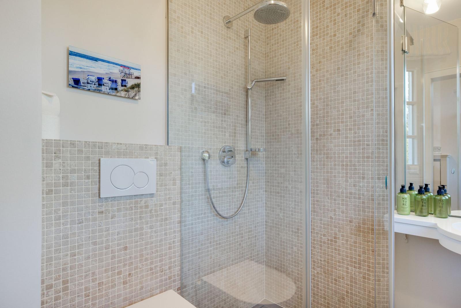 Dusche mit Glaswand, Mosaikfliesen und Duschbrause. Toilette und Waschbecken mit Flaschen.
