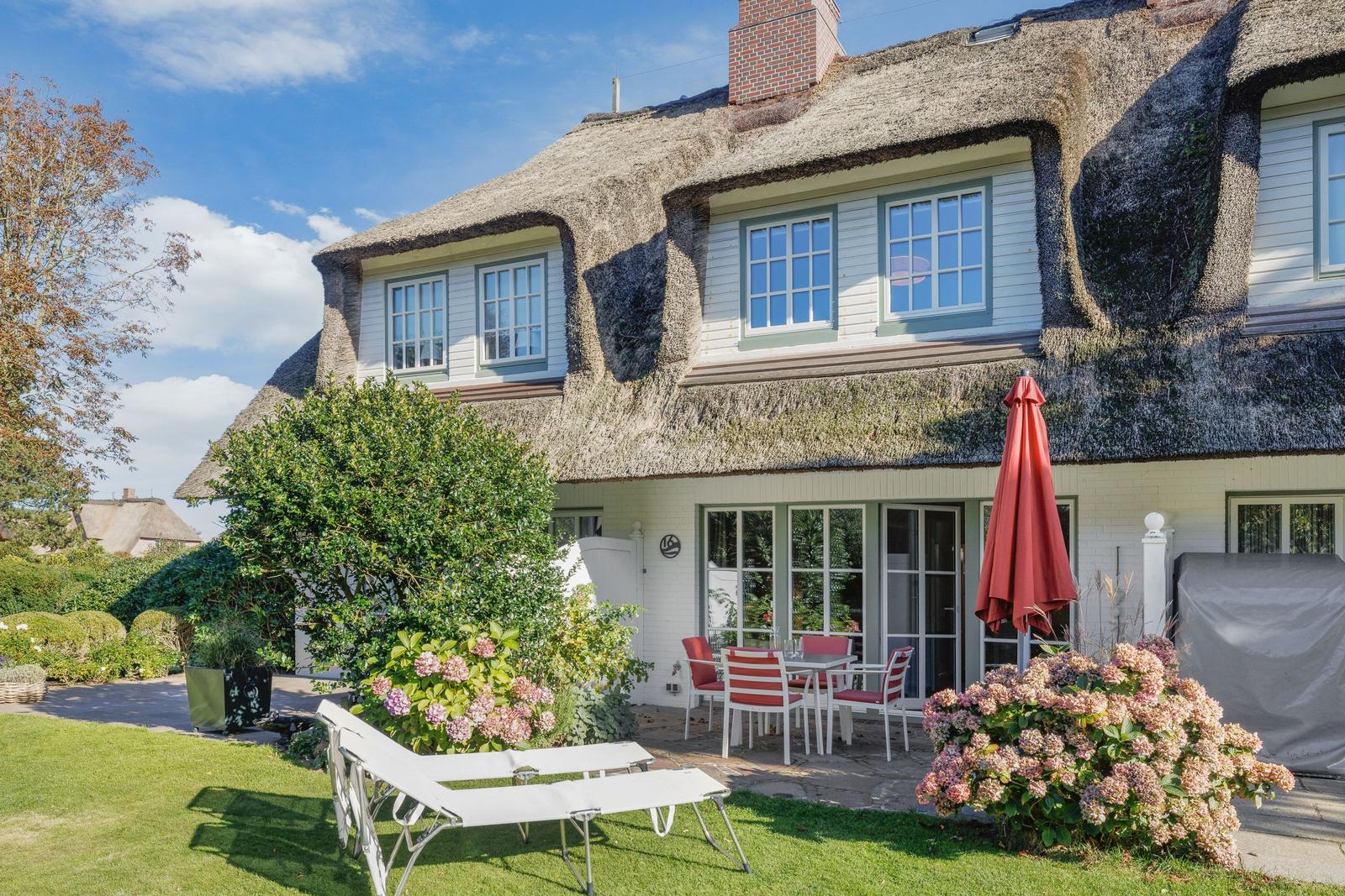 Haus mit Strohdach, Terrasse mit Tisch und Stühlen, Liegestühle auf Rasen, Blumenbeete und Sonnenschirm.