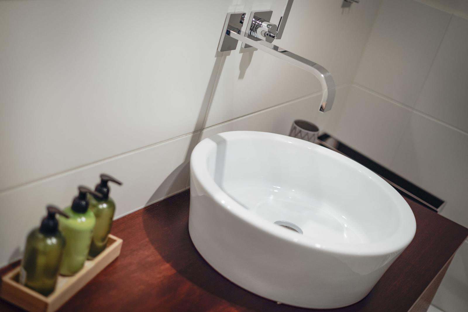 Waschbecken auf Holzunterschrank mit modernem Wasserhahn und grünen Flüssigseifenflaschen.