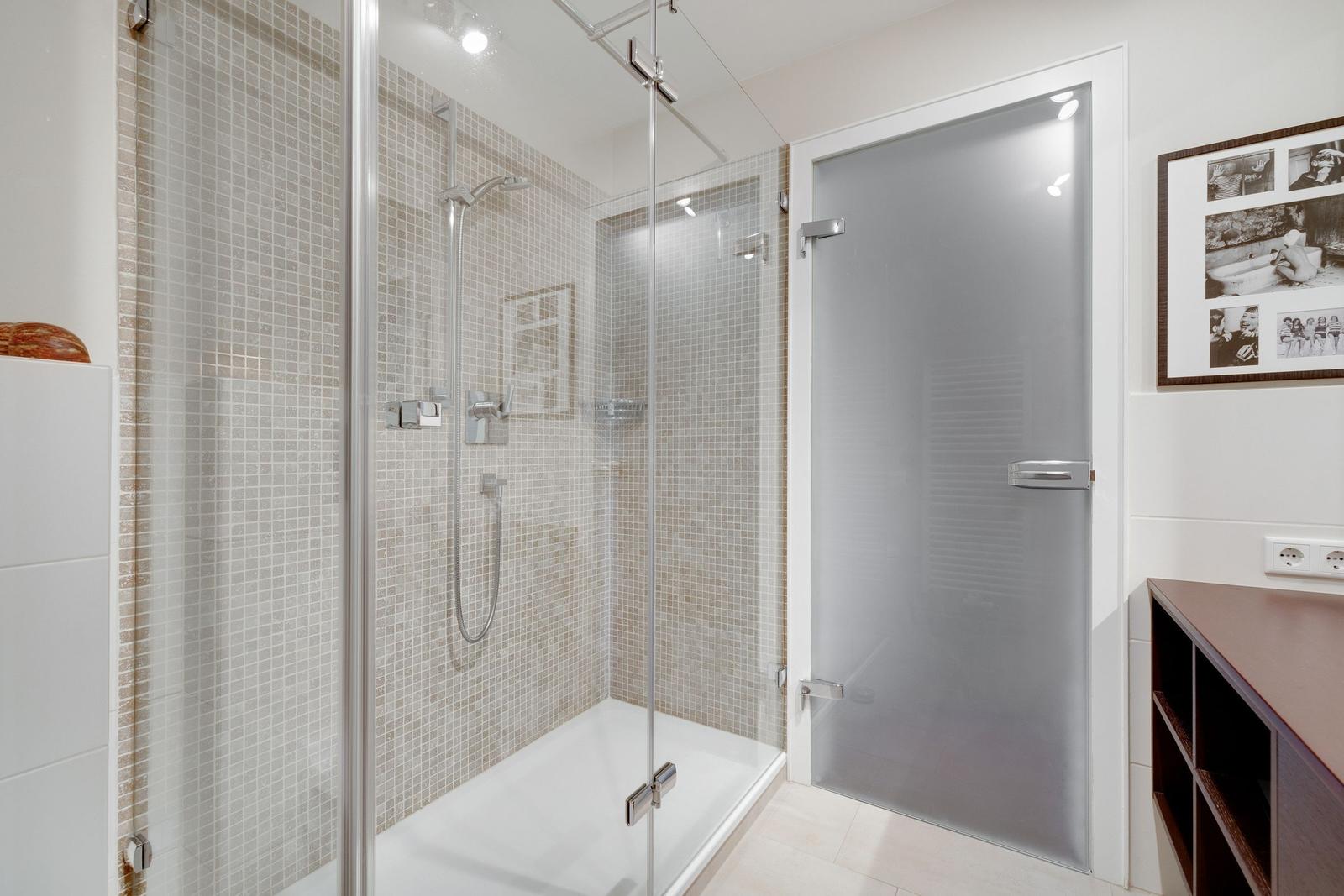 Dusche mit Glaswand und Mosaikfliesen, Nebenraum mit Schrank und Bild an der Wand.