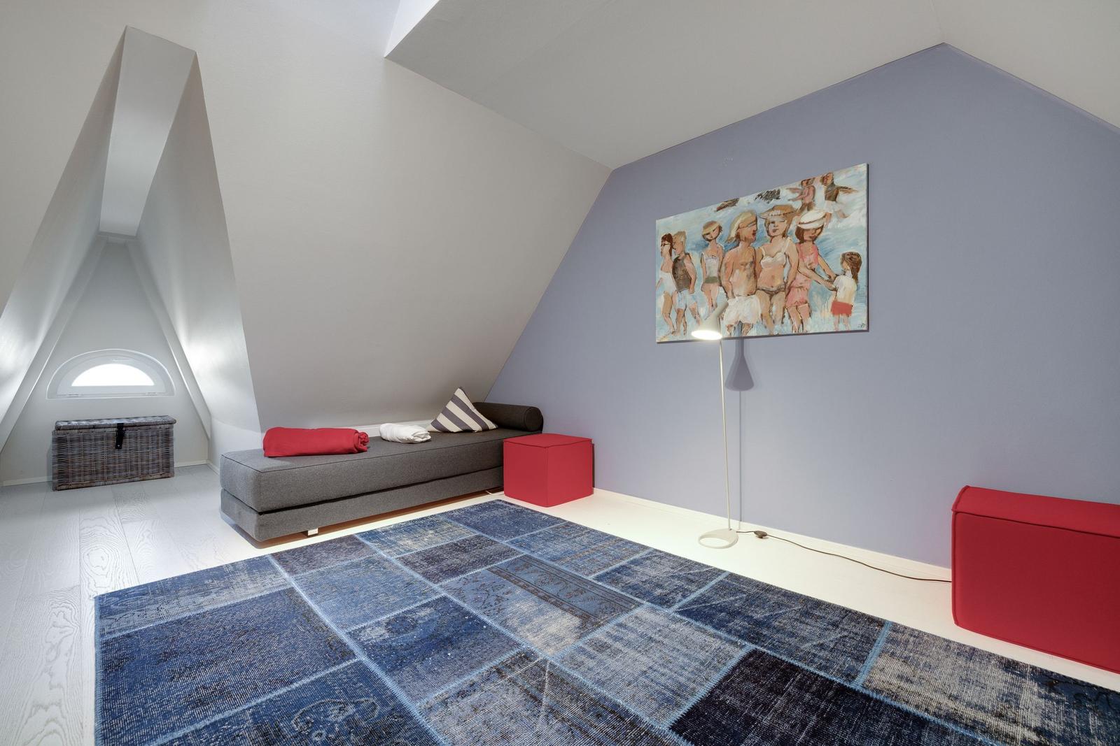 Dachgeschosszimmer mit grauer Couch, rotem Sitzkissen und blauem Teppich.