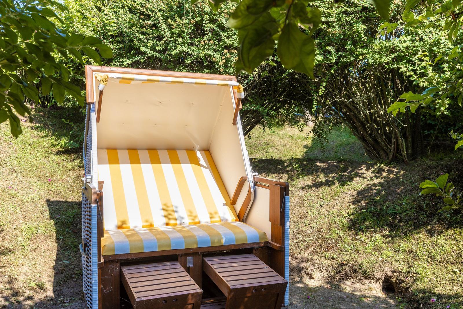 Holzliege mit gelb-weißem Streifenbezug im Garten unter Bäumen.