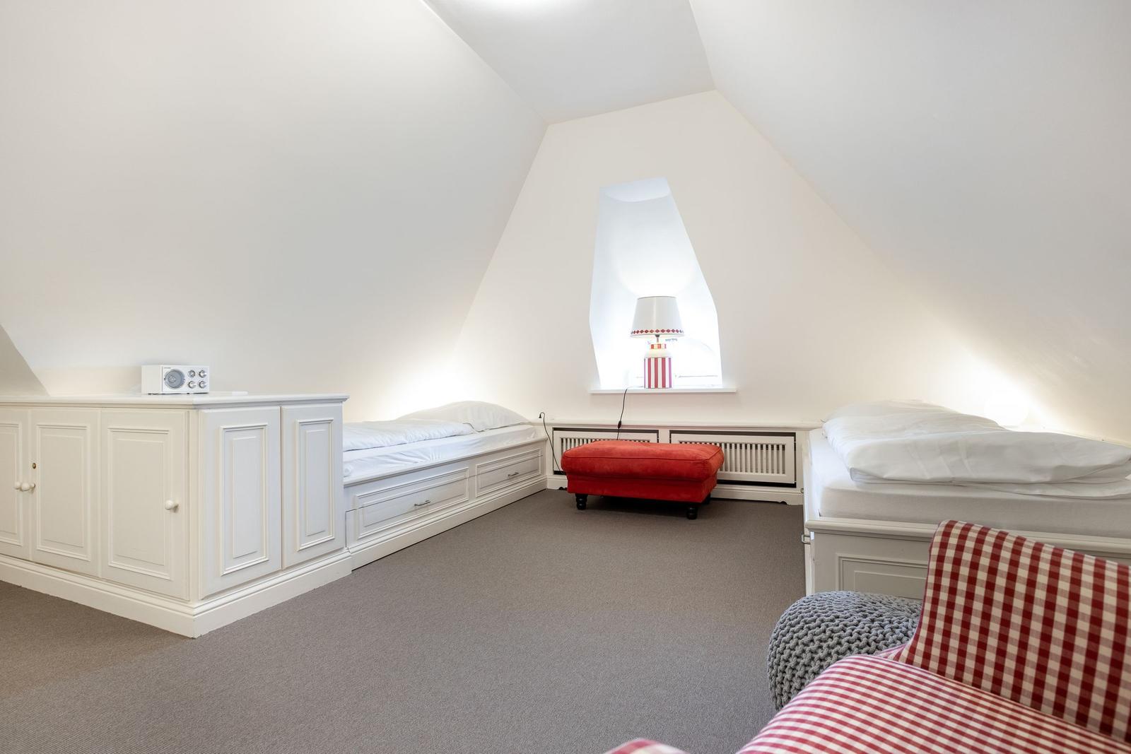 Zimmer mit zwei Betten, Schrank, rotem Sitzkissen und Fenster unter Dachschrägen.