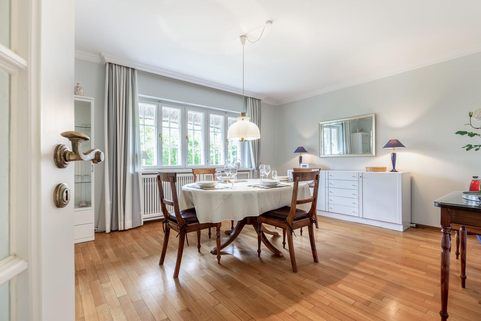 Dining room with wooden floor, table and chairs, windows and mirror on the wall.
