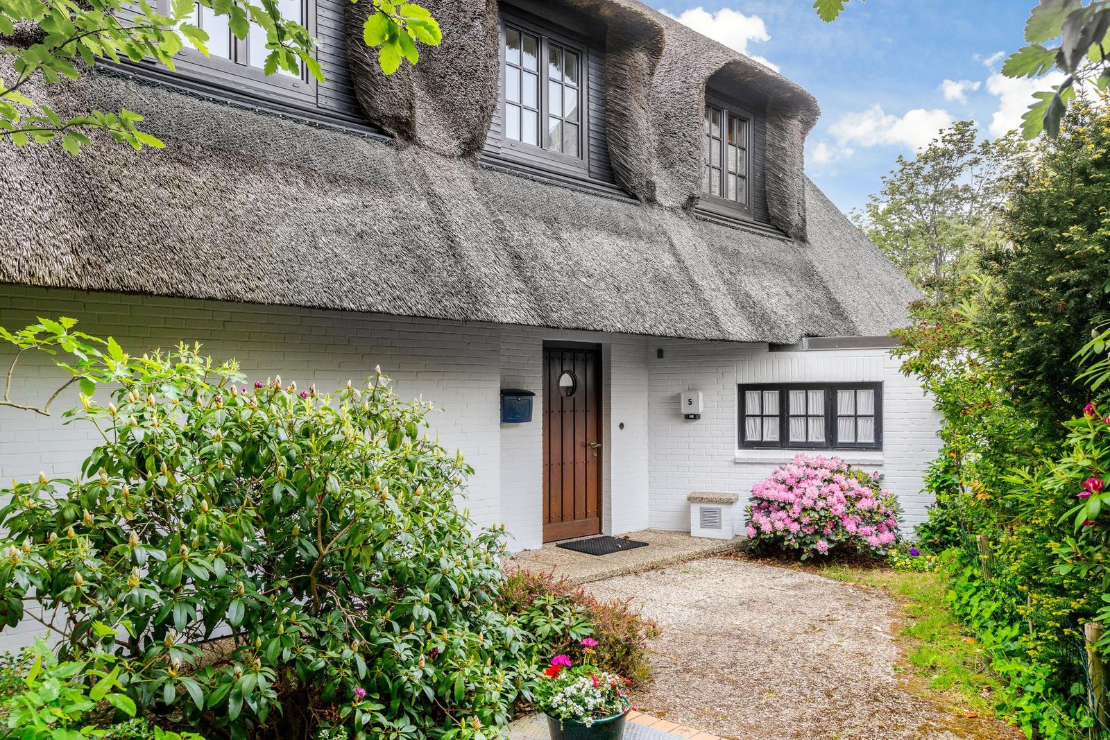 House with thatched roof, white facade, and blooming plants in the front garden.