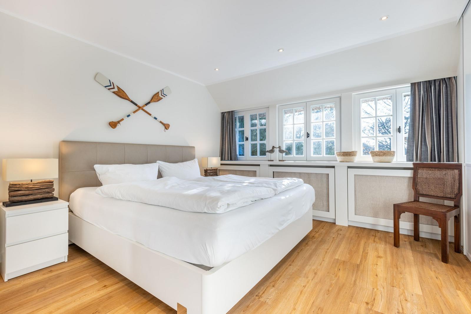 Schlafzimmer mit Bett, Nachttisch, Stuhl und Fenstern.