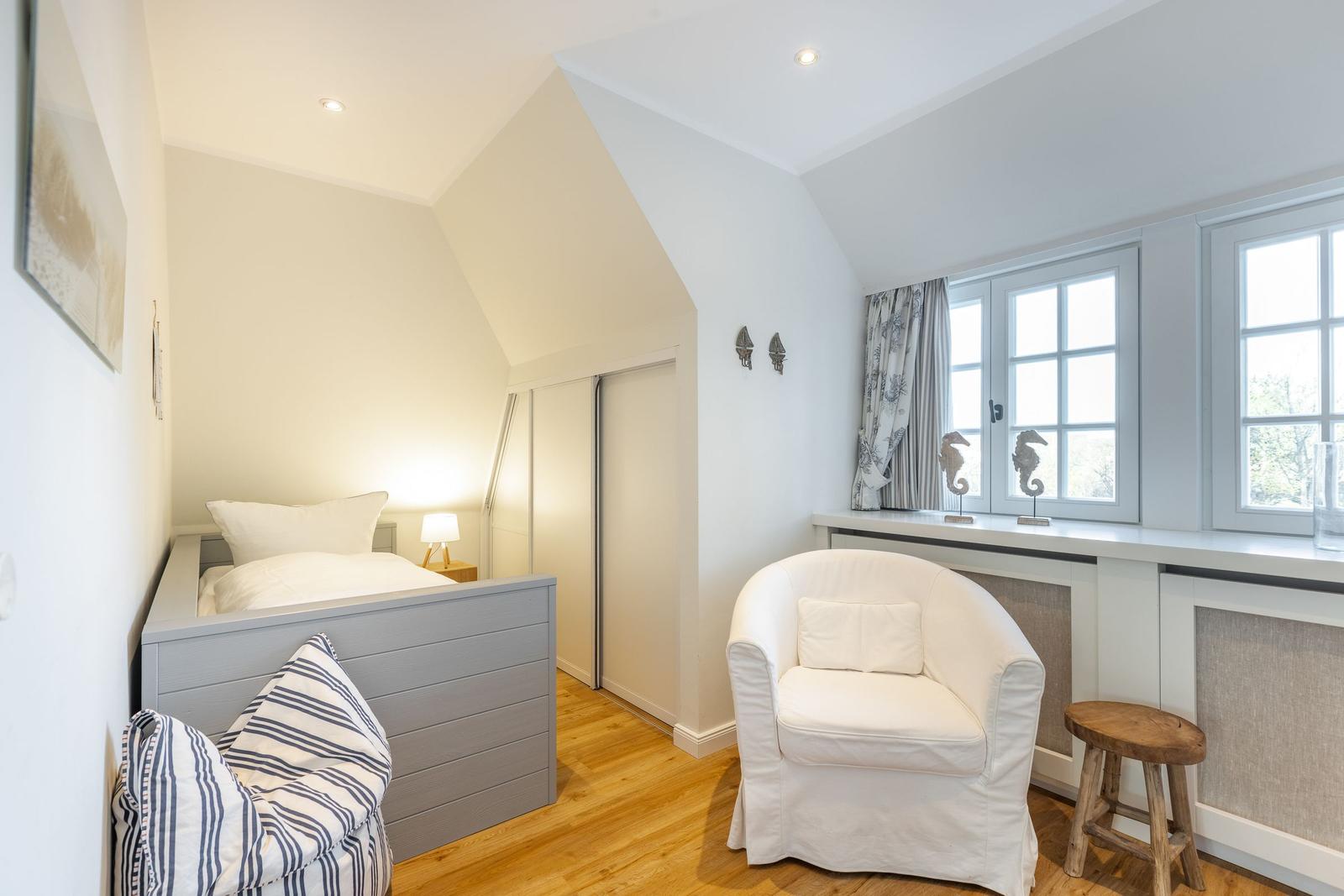Schlafzimmer mit Bett, Sessel und Fenster. Holzboden und weiße Wände.