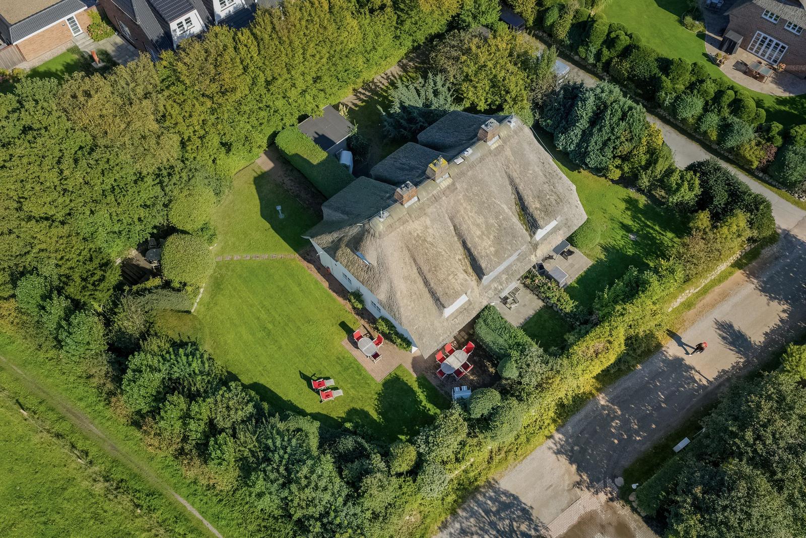House with thatched roof, surrounded by lawn and trees. Outdoor area with red garden furniture.