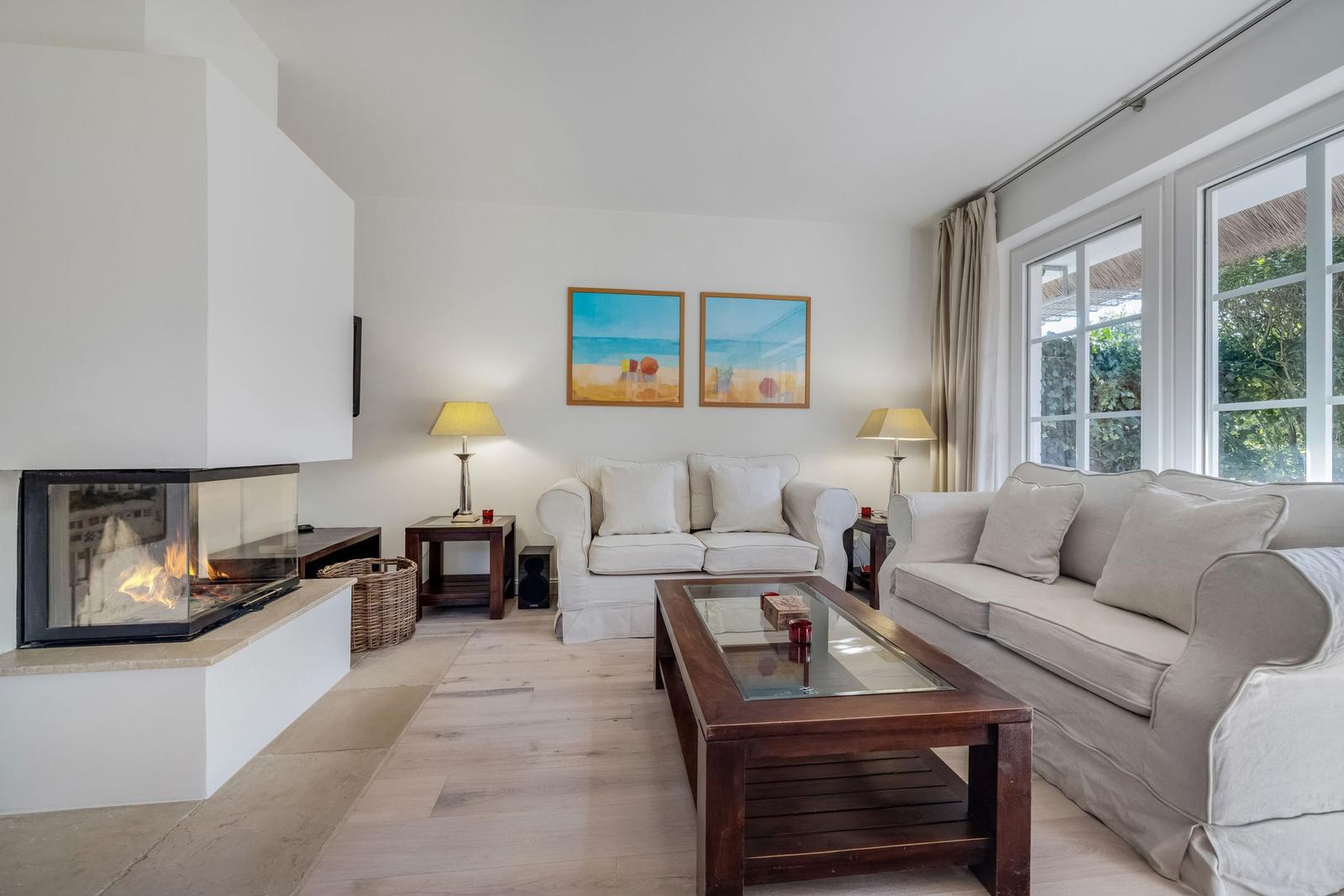 Living room with fireplace, two sofas, and glass-top table. Windows show garden.