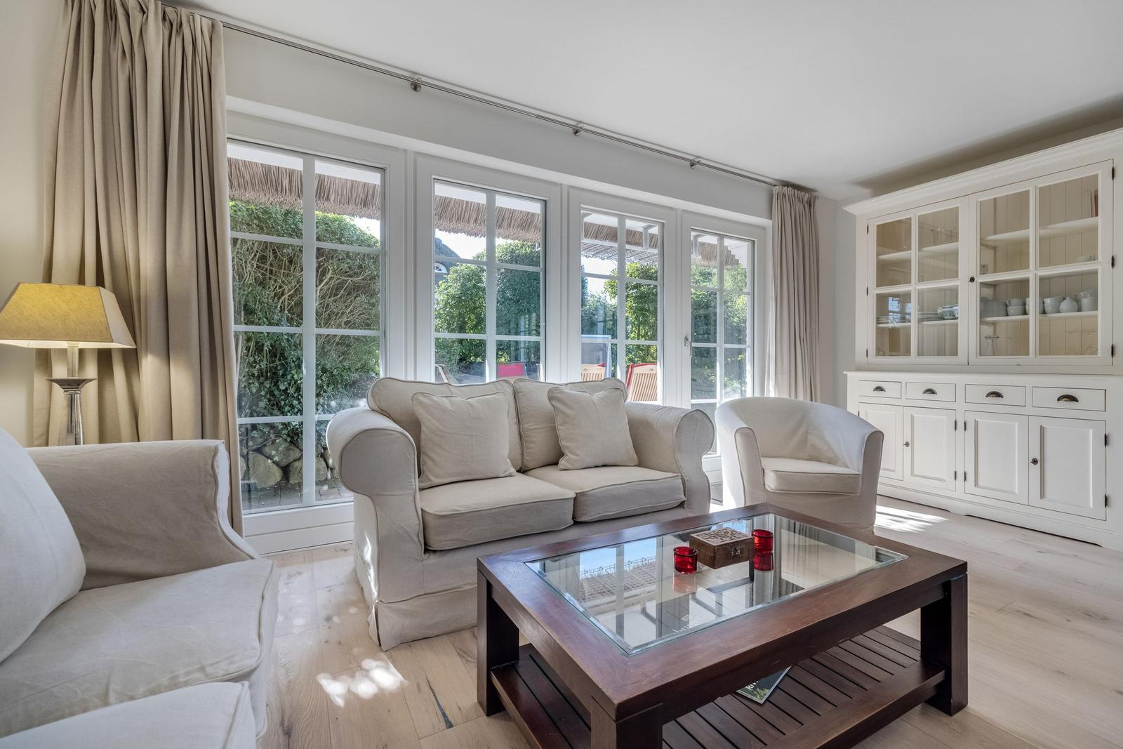 Living room with sofa, armchairs, and glass-top table. Large windows and white cabinets.