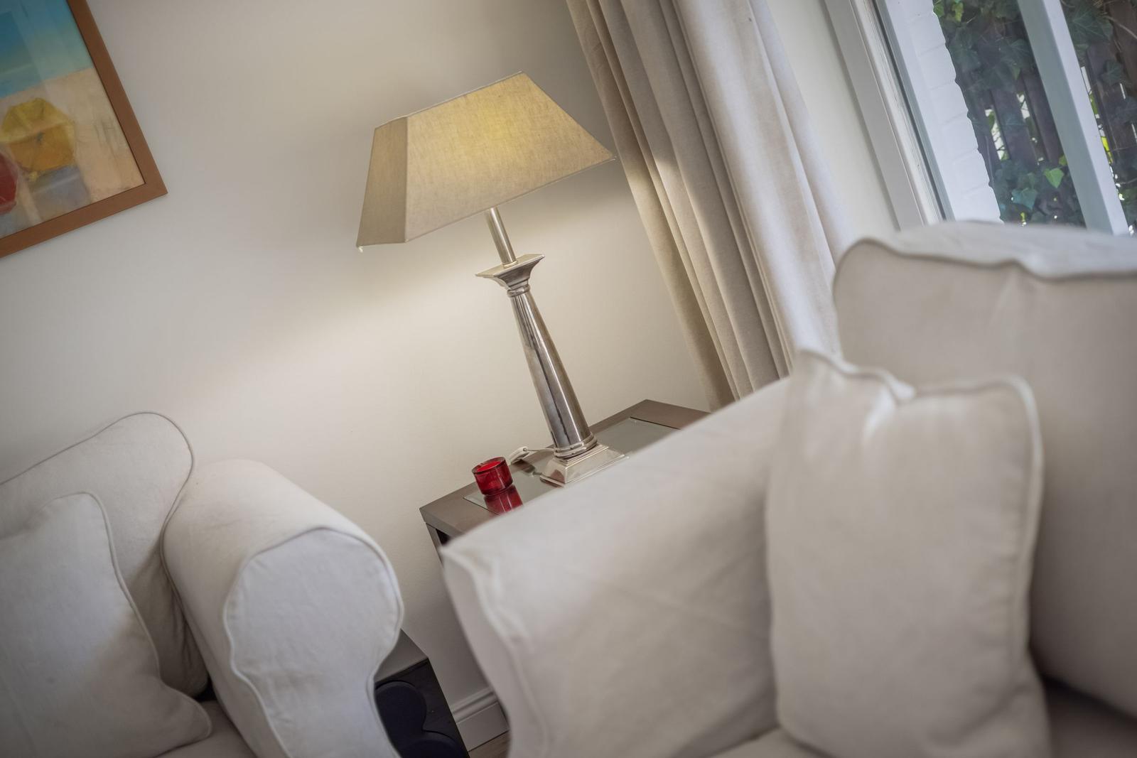A white armchair next to a nightstand with lamp and red glass.