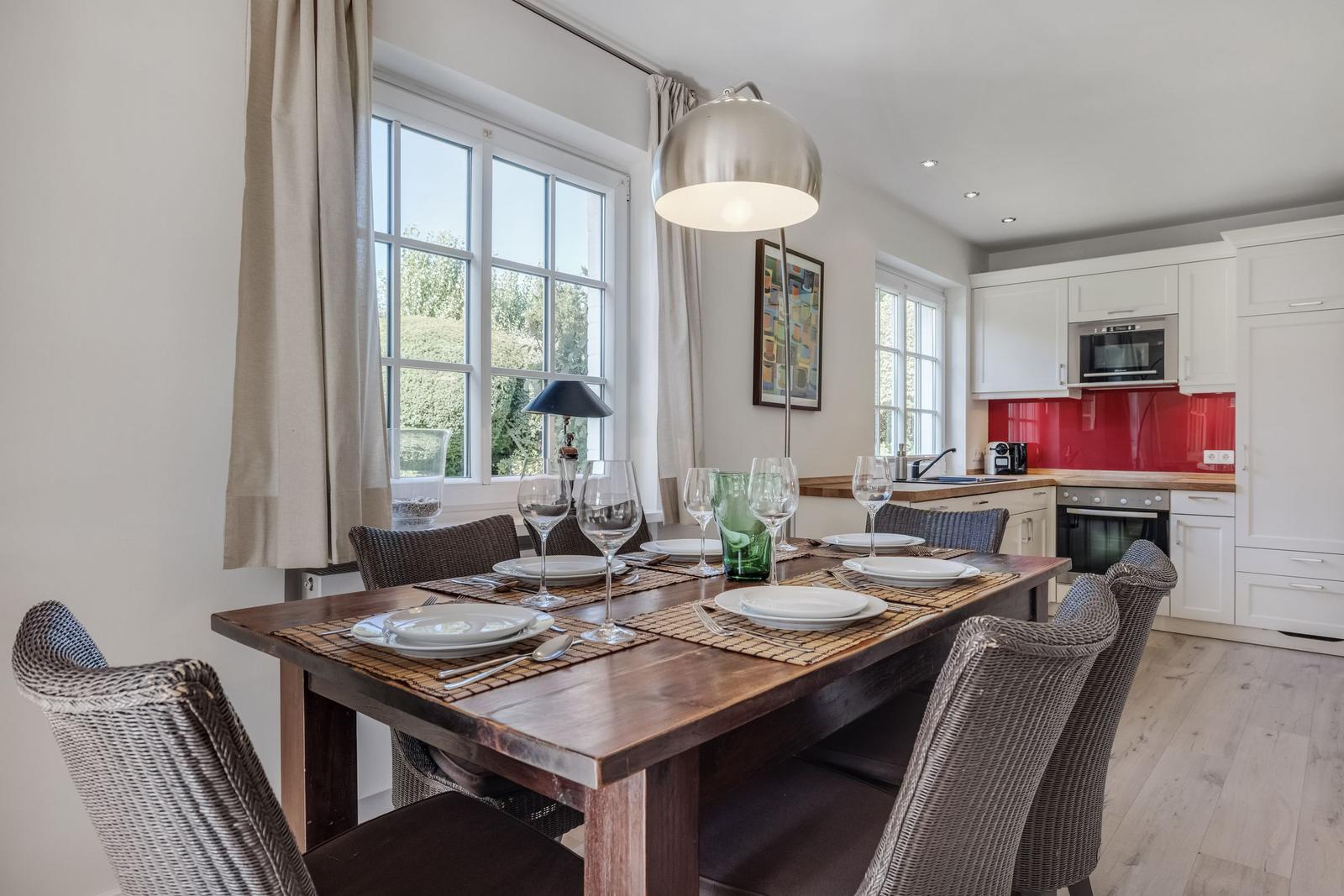 Dining room with table, chairs, and windows. Kitchen with red backsplash and white cabinets.
