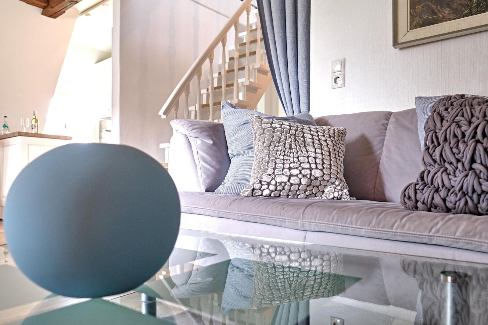 Ein Sofa mit Kissen und Decke vor einem Glas-Tisch. Hintergrund: Treppe und Fenster.