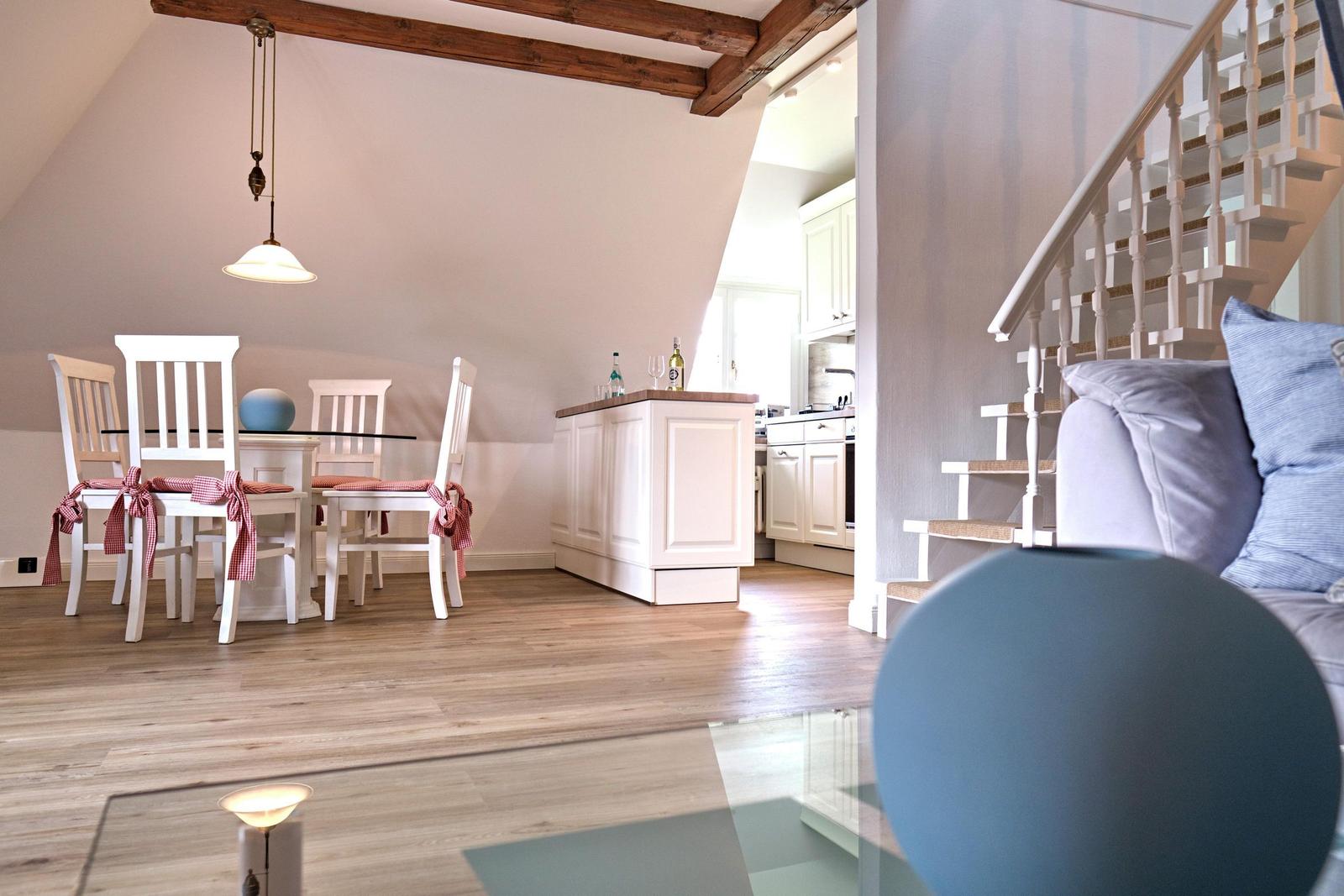 Essbereich mit Tisch, Stühlen und Küchenbereich. Holzdielenboden und Treppe sichtbar.