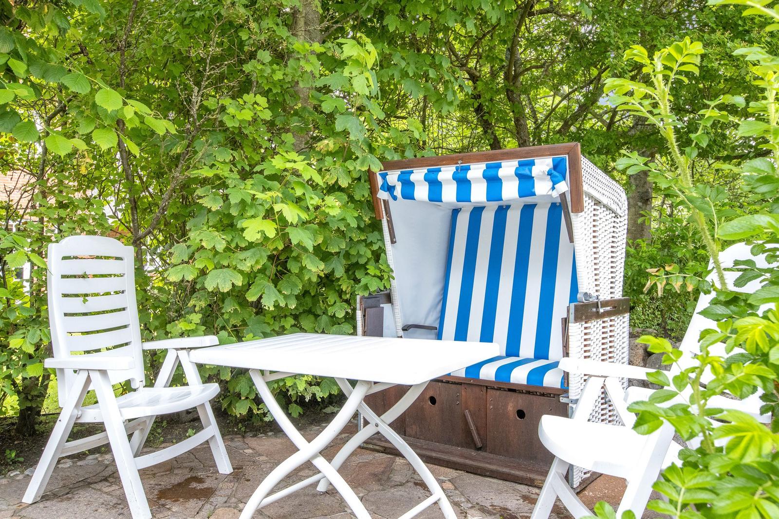 Behind green bushes, a white terrace with a blue-and-white striped beach chair and white chairs.