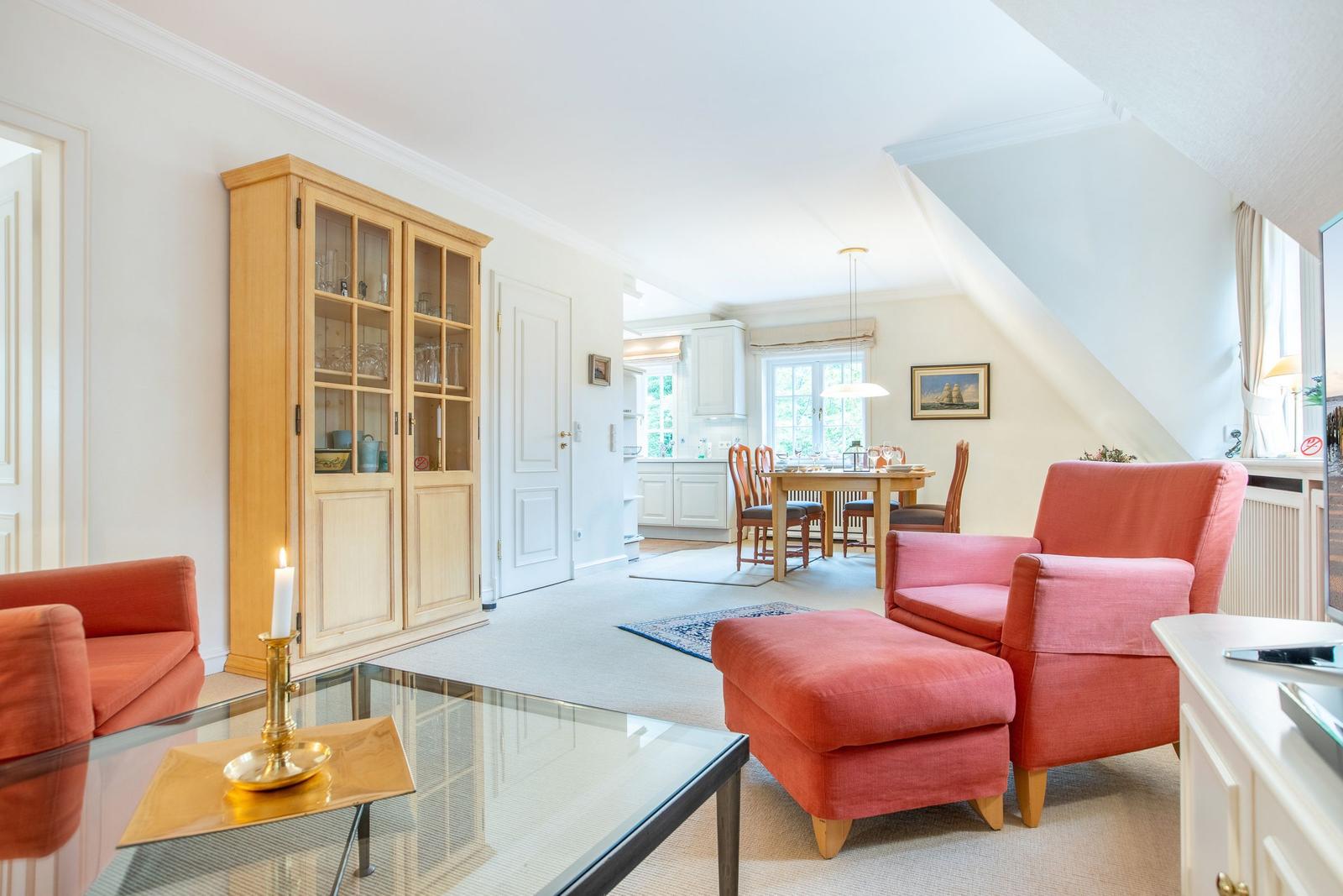 Living room with red armchairs, glass table, and wooden cabinet. Kitchen and dining area in background.