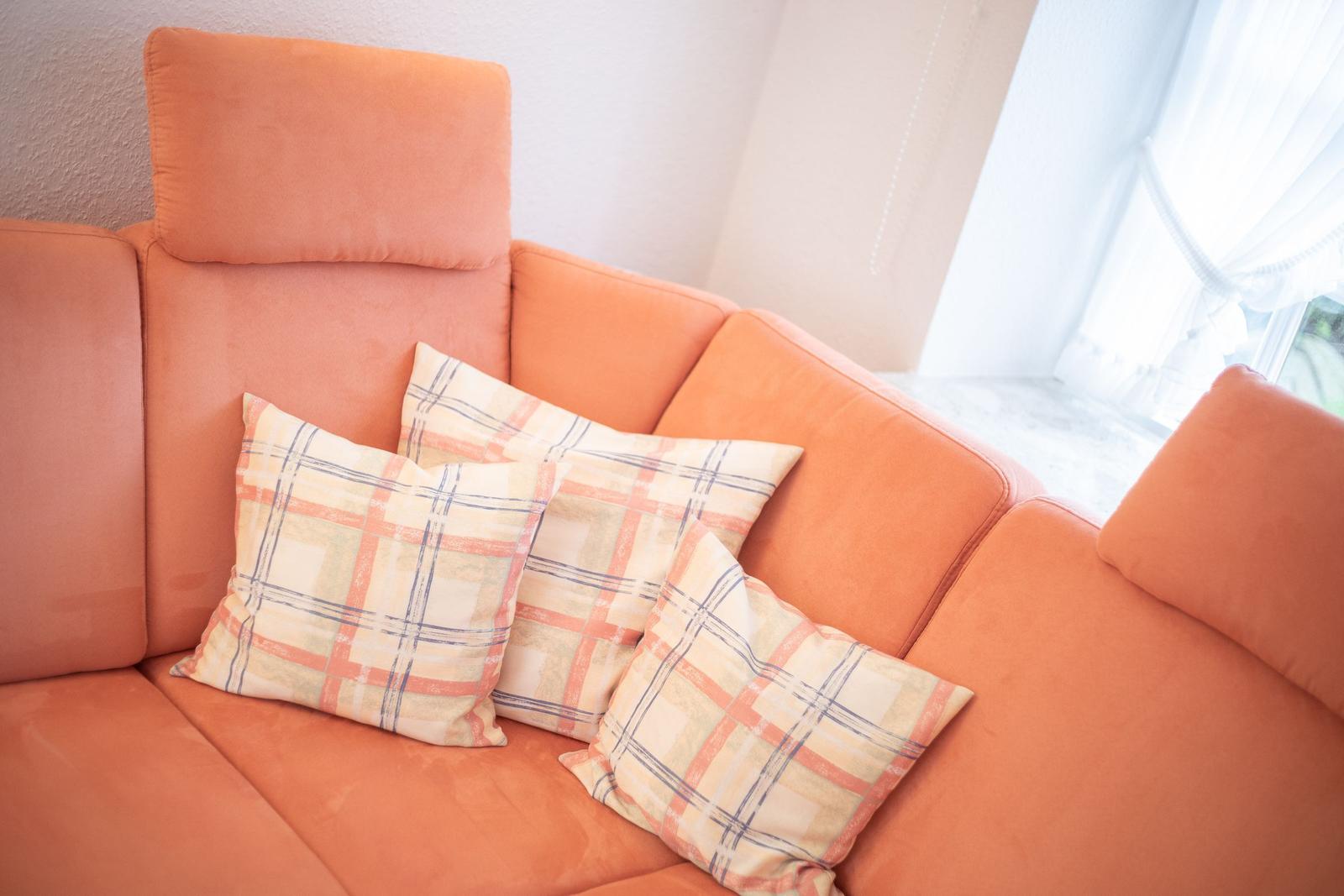 Orangefarbener Sofa mit drei karierten Kissen und Fenster im Hintergrund.