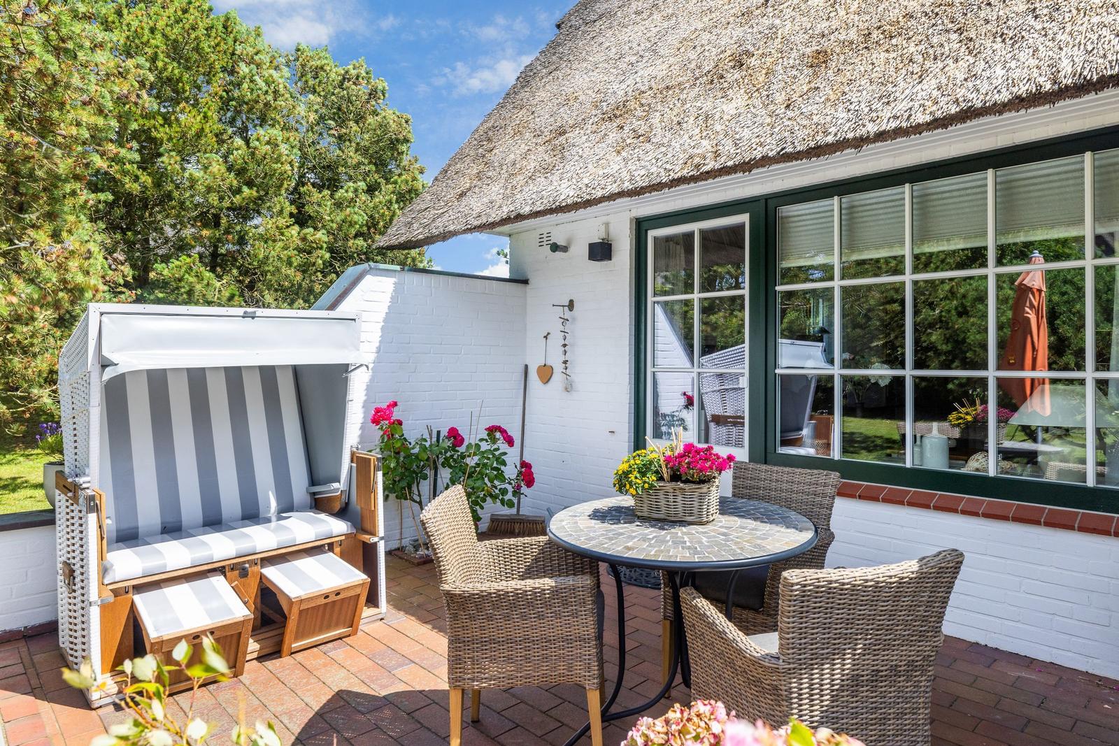Terrasse mit Rattanmöbeln, Strandkorb und Blumen vor einem Haus mit Strohdach.