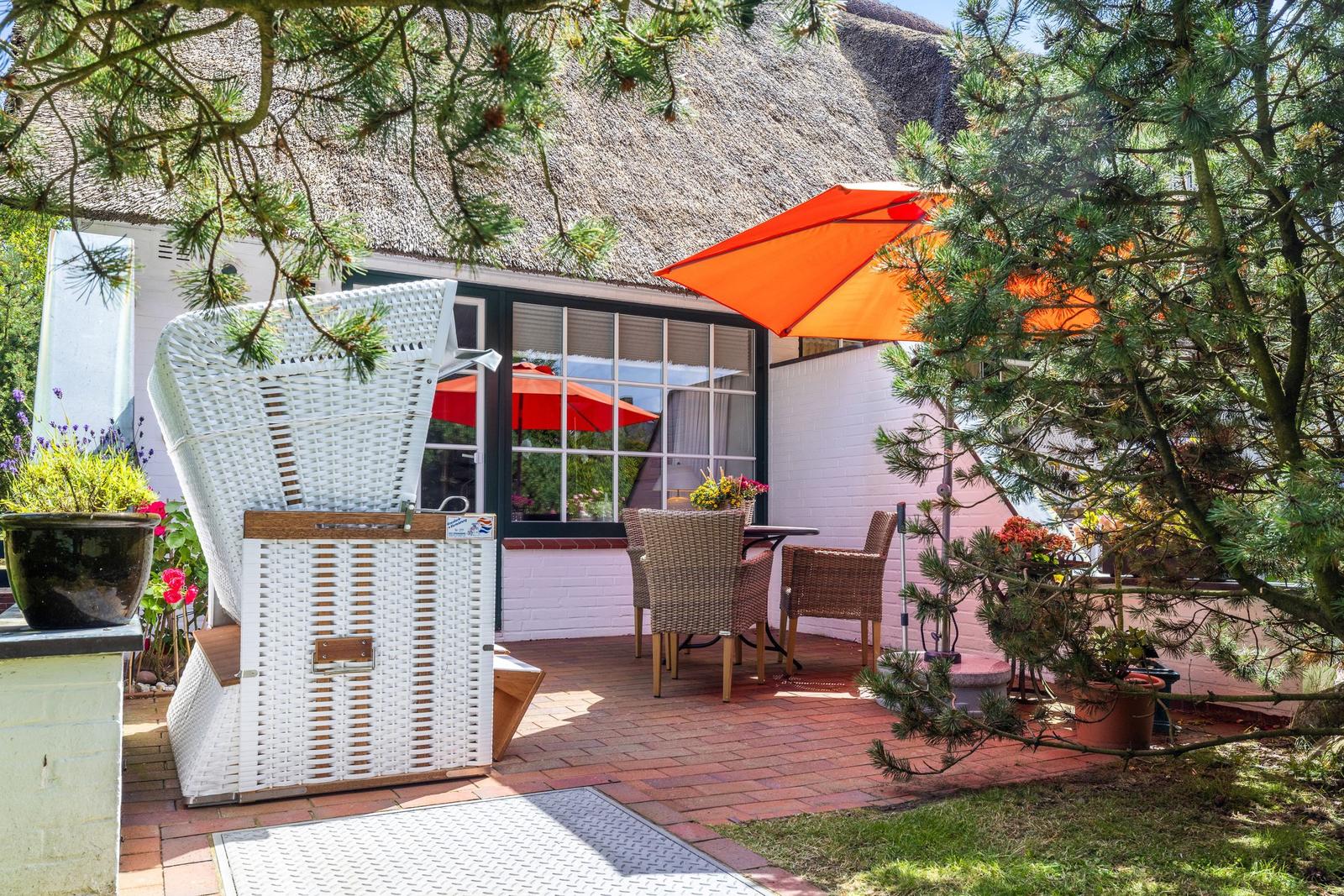 Terrasse mit orangefarbenem Sonnenschirm und weißem Liegestuhl vor einem Haus mit Strohdach.