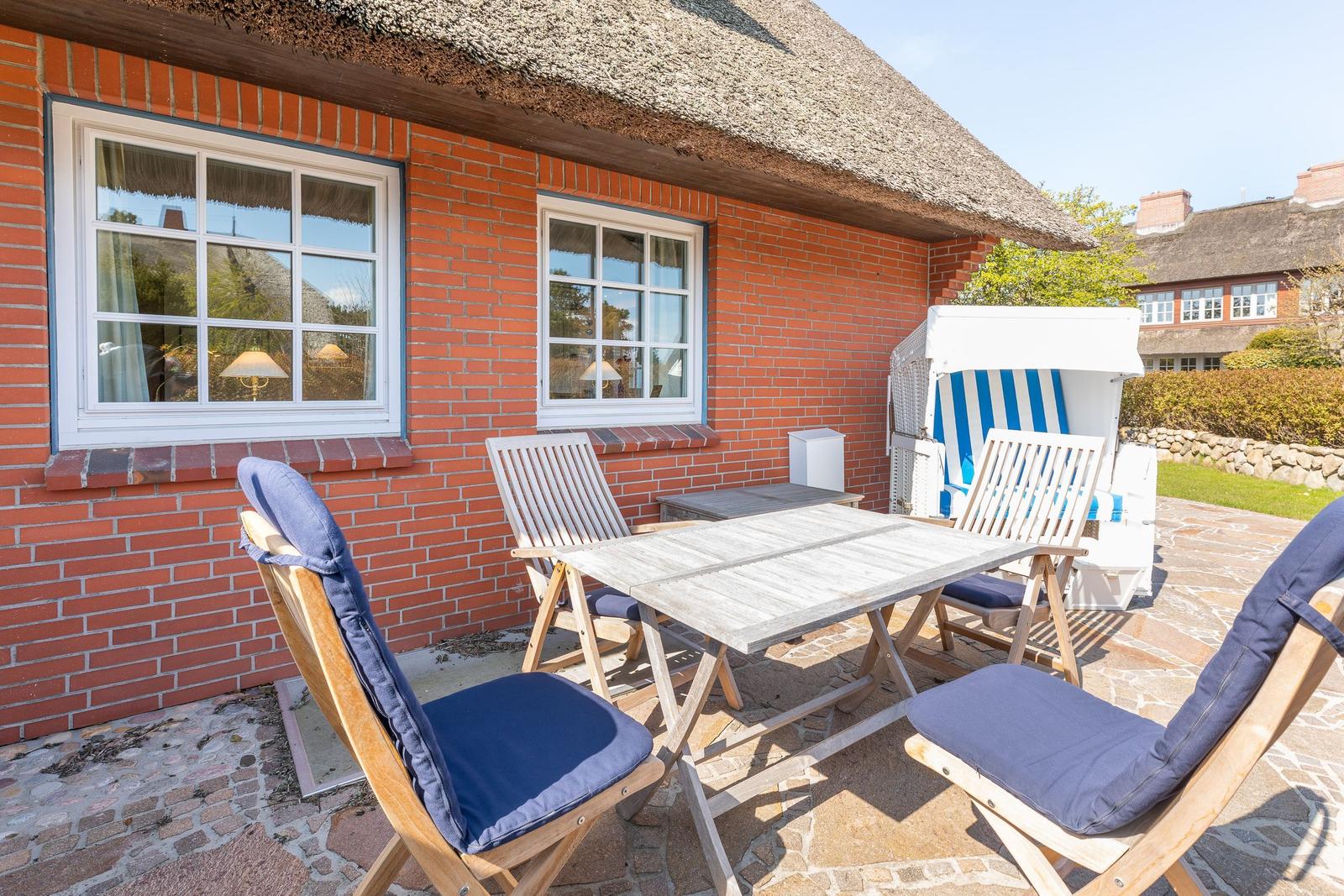 Terrace with table, chairs, and beach chair in front of red brick house with thatched roof.