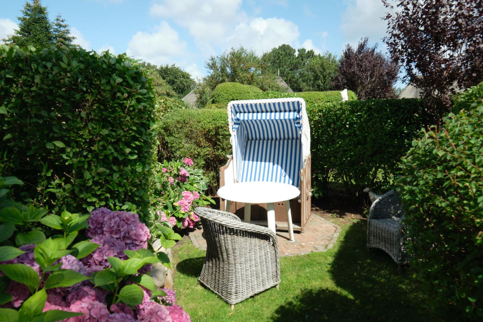 Garten mit Strandkorb, Tisch und Stühlen umgeben von Sträuchern und Blumen.