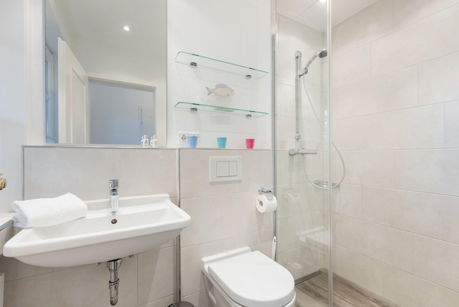 Badezimmer mit Dusche, Waschbecken und Toilette. Glasregale und Spiegel sichtbar.
