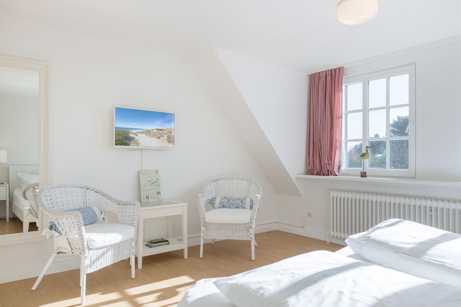 Schlafzimmer mit Dachschräge, zwei weißen Stühlen und Fenster mit rotem Vorhang.