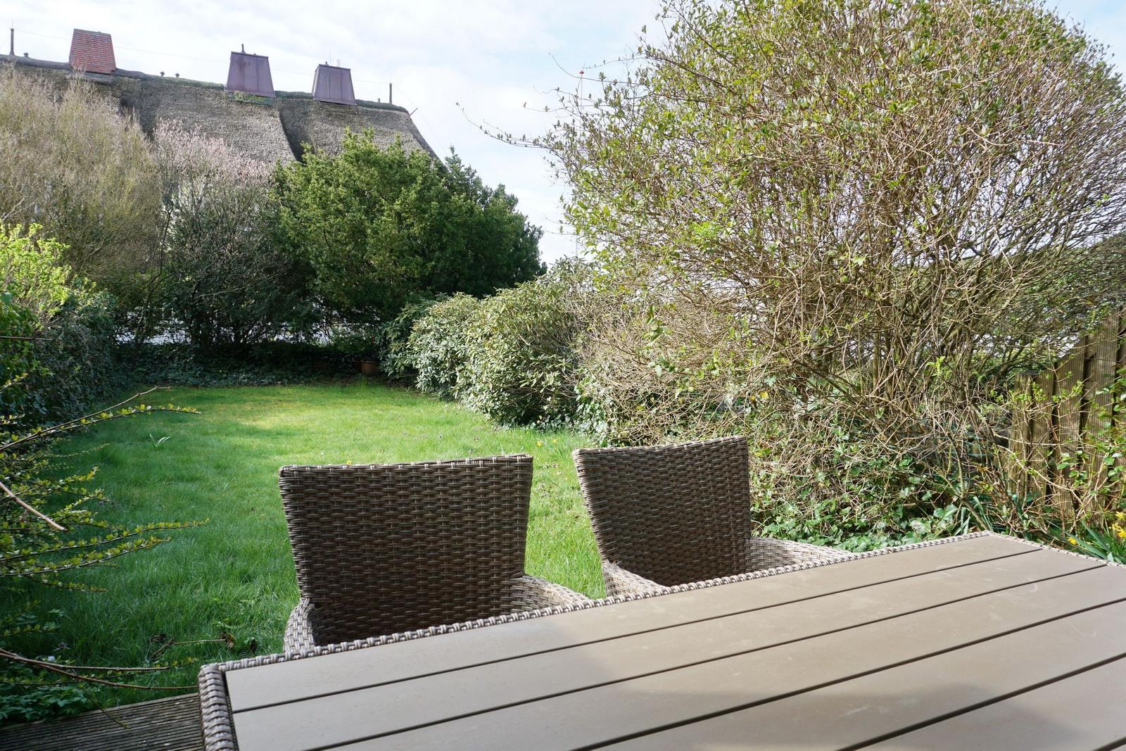 Terrasse mit Tisch und zwei Stühlen. Grünfläche und Bäume im Hintergrund.