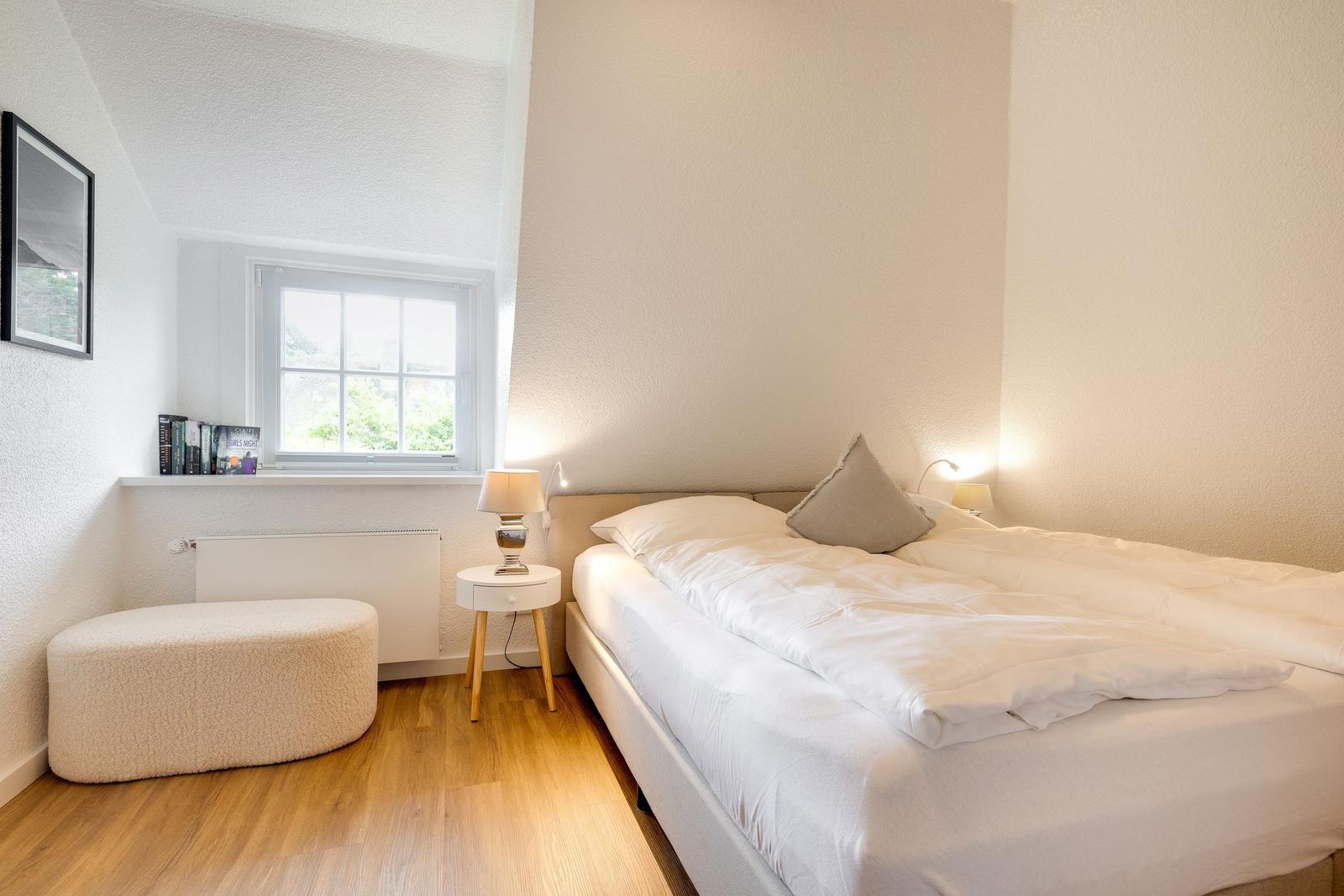 Schlafzimmer mit Bett, Nachttisch und Fenster. Holzboden und Wandheizung.
