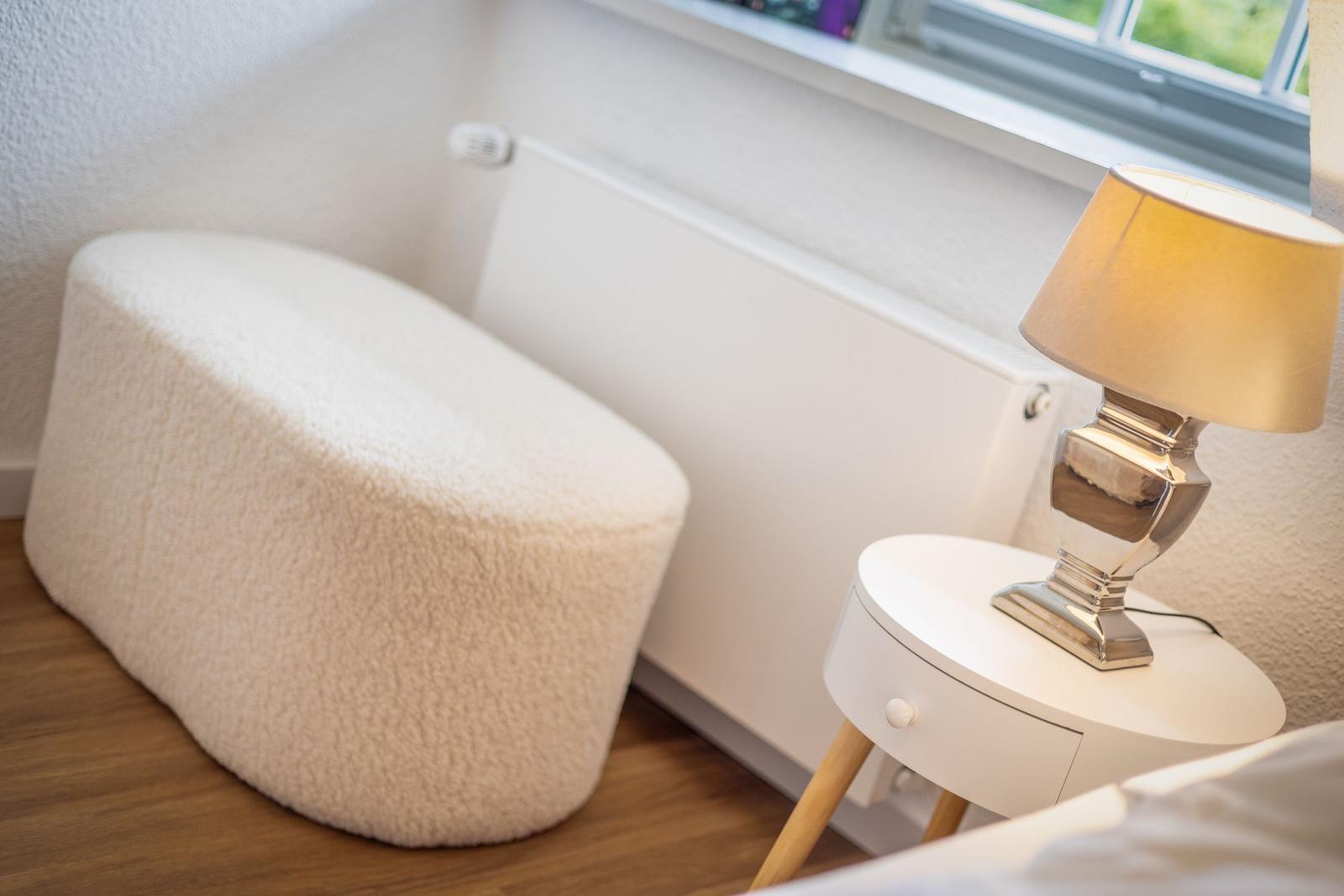 Weißer, flauschiger Sitzhocker neben Nachttisch mit Lampe.
