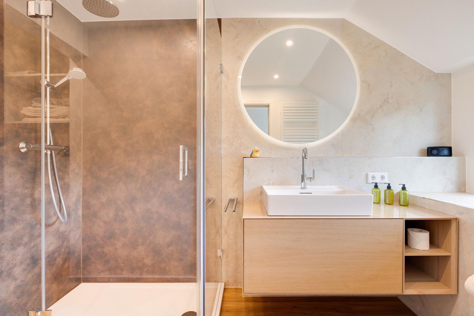 Dusche mit Glaswand, Waschbecken mit runden Spiegel und Holzunterschrank.