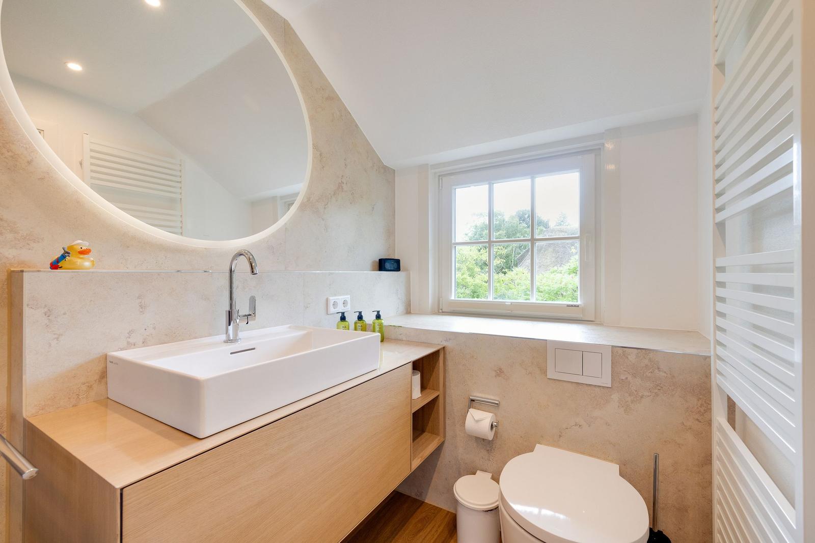 Badezimmer mit Waschtisch, Spiegel und Fenster. Toilette und Heizung sind vorhanden.