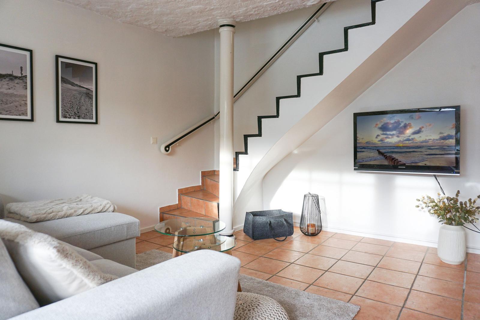 Wohnzimmer mit Sofa, Glas-Tisch und Treppenhaus. Fernseher an Wand montiert.