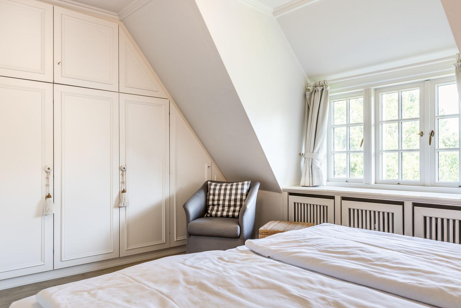 Schlafzimmer mit Dachschrägen, weißem Schrank und Fenster mit Blick auf Bäume.
