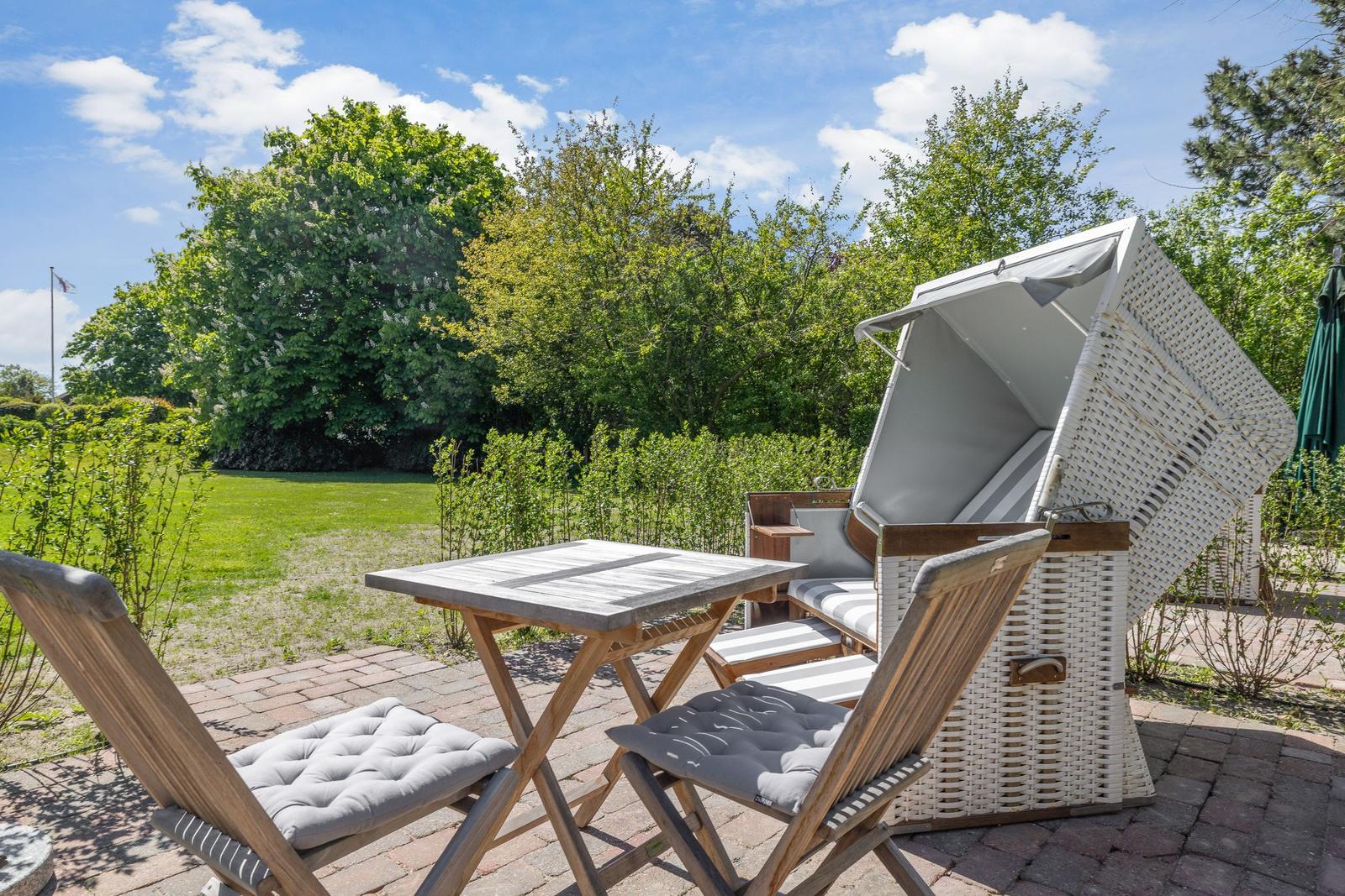 Terrasse mit Tisch, Stühlen und Strandkorb. Hintergrund: grüner Garten und Bäume.