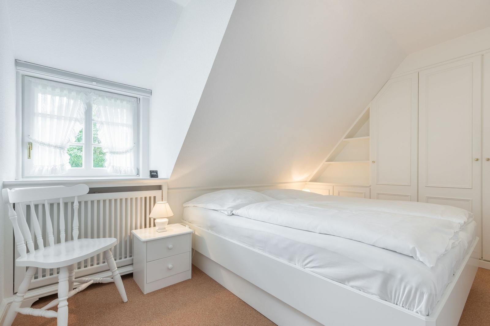 Schlafzimmer mit weißem Bett, Nachttisch und Stuhl unter Dachschrägen.